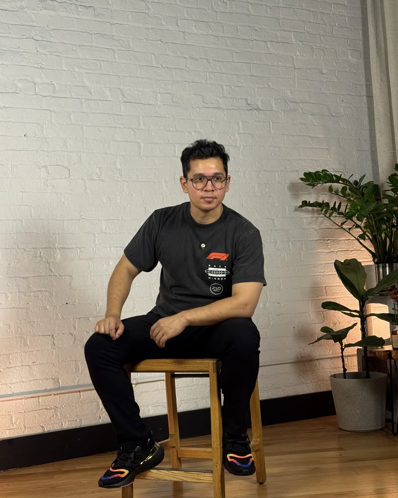 Jenesh sitting on a wooden stool against a white brick wall